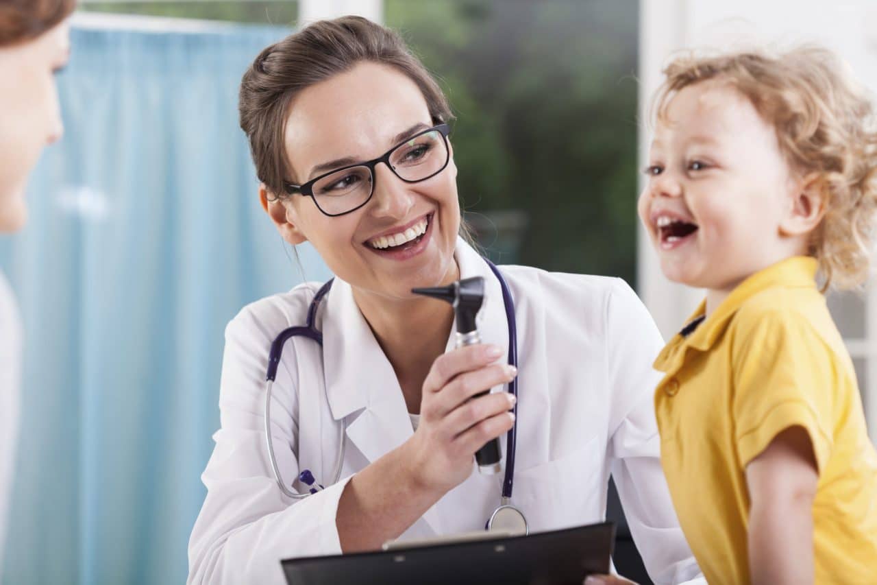 An audiologist showing an otoscope to a child who's laughing at another adult