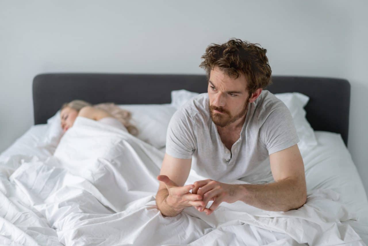 A person with rumpled hair sitting up in bed while their partner sleeps
