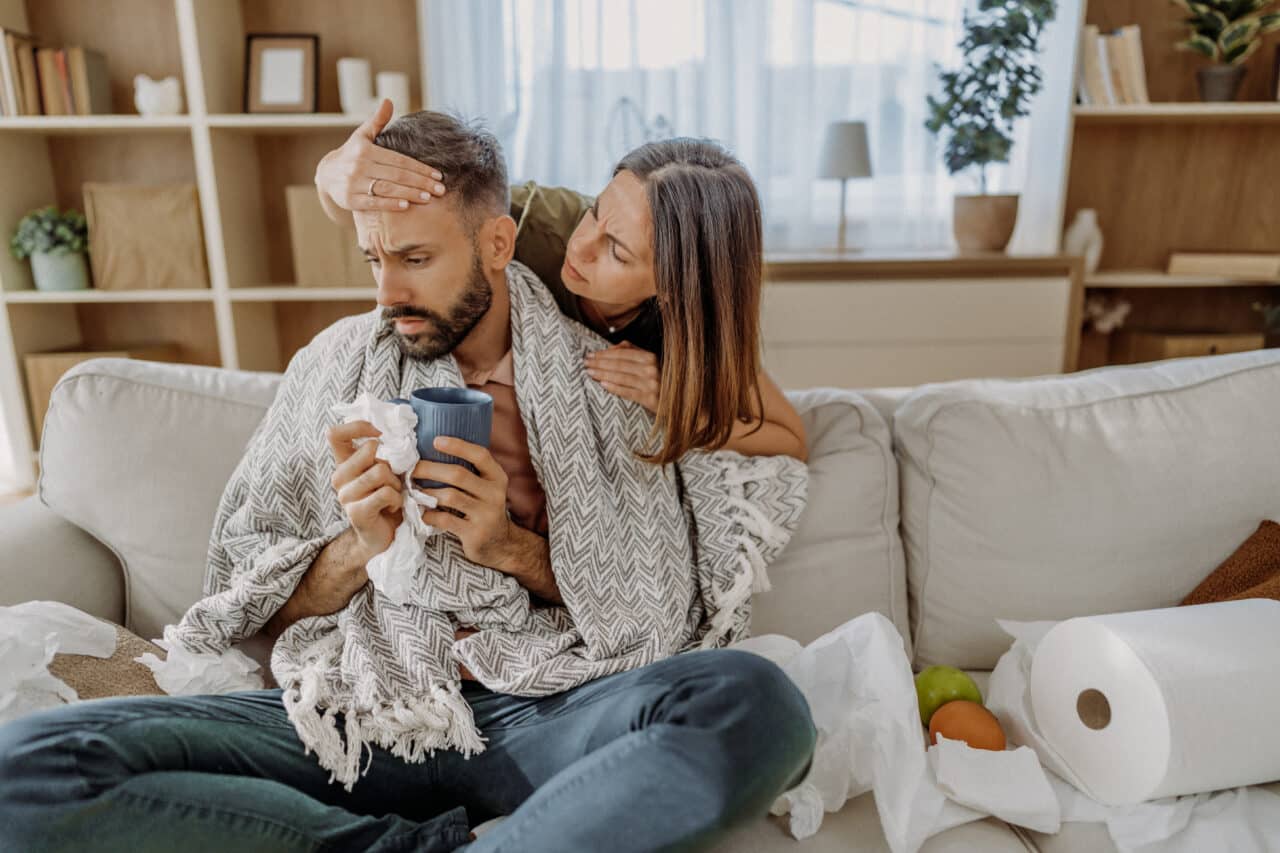 Man with cold-like symptoms sits on the couch. A woman feels his forehead for a fever to determine if it's allergies or a virus.