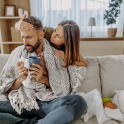 Man with cold-like symptoms sits on the couch. A woman feels his forehead for a fever to determine if it's allergies or a virus.