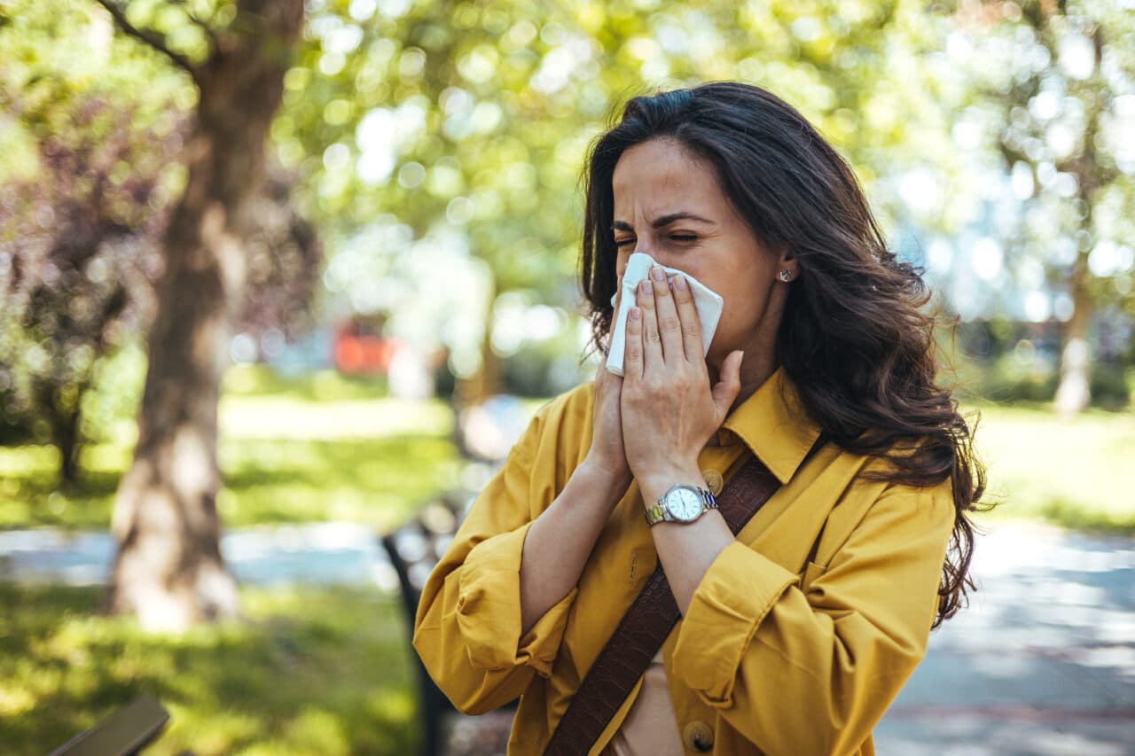Woman sneezing, blowing her nose, suffering from allergies while taking a walk in the park.