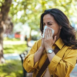Woman sneezing, blowing her nose, suffering from allergies while taking a walk in the park.